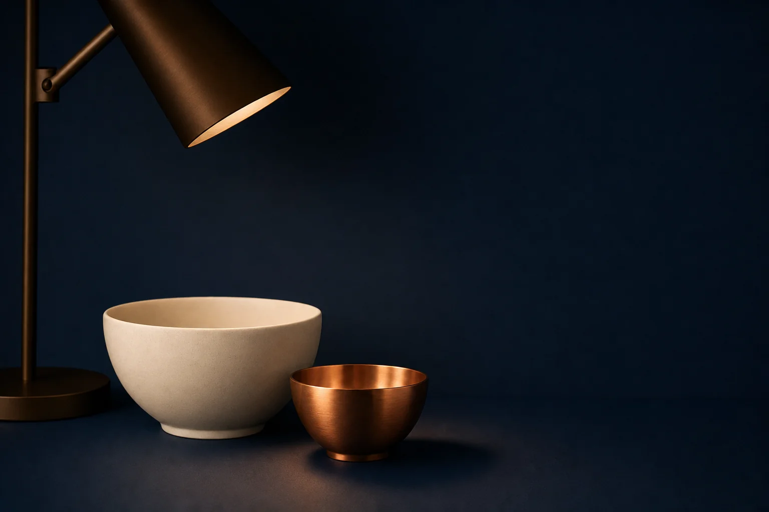 A large ivory ceramic bowl and a smaller copper bowl side by side on a navy desk beneath a bronze desk lamp