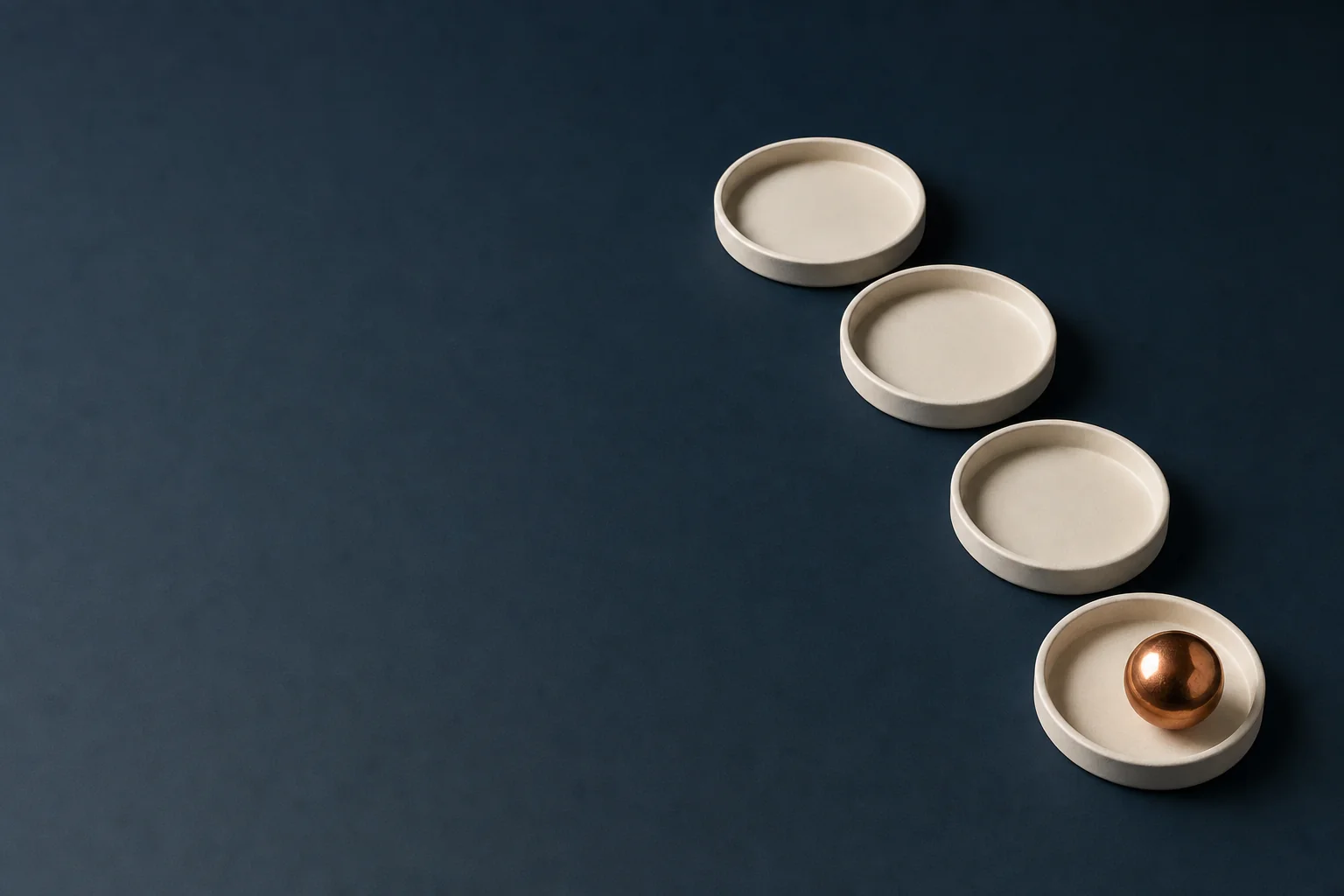 Four shallow round ivory dishes in a receding diagonal on a navy surface, the nearest one holding a polished copper sphere