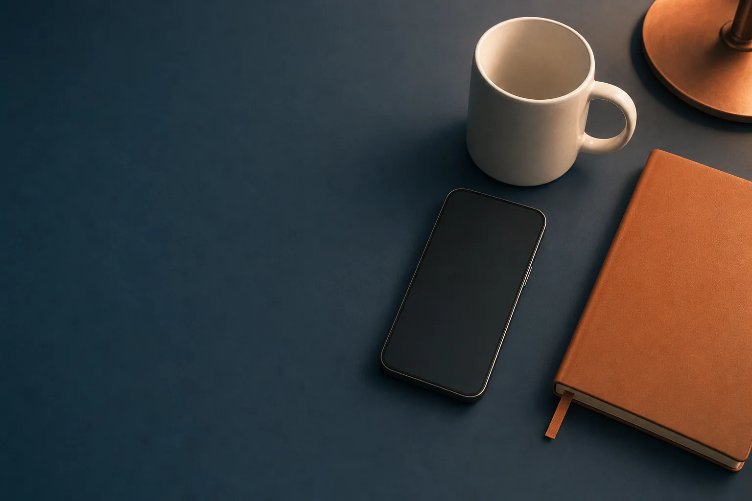 A smartphone with a blank screen on a dark navy desk beside a copper-colored notebook and a white ceramic mug