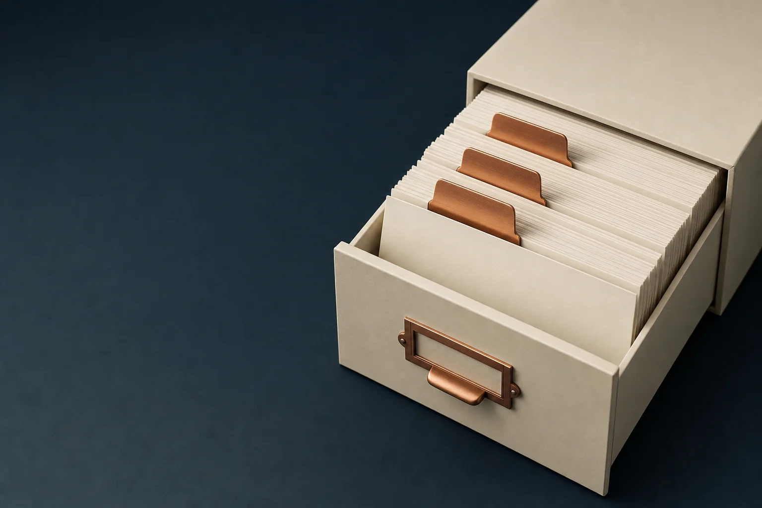A shallow ivory drawer of upright blank index cards separated by three copper divider tabs on a navy surface