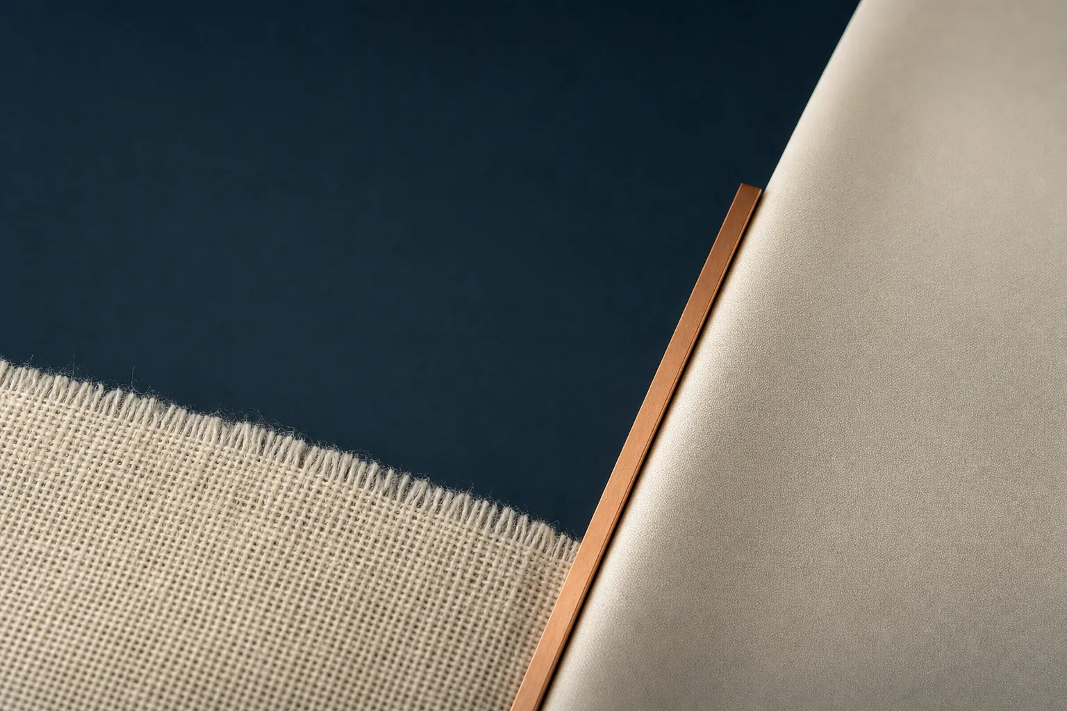 A coarse ivory open-weave cloth with loose fibres along its frayed edge at lower left and a smooth pale ivory sheet at upper right, separated by an open navy field and a slim copper bar running along the diagonal between them.